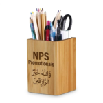 Square bamboo pen holder on a white background, filled with pens and pencils, featuring custom engraved text and Arabic calligraphy on the front panel.
