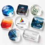 A collection of clear custom acrylic paperweights in round, square, and rectangular shapes, featuring full-color printed corporate logos and nature designs.