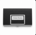 A top-down view of a black textured faux-leather business card holder with polished metal trim, featuring a central metal plaque laser-engraved with the name "Abdul Rafay". The holder is closed and centered on a pure white background.