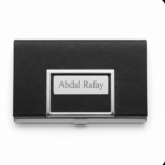 A top-down view of a black textured faux-leather business card holder with polished metal trim, featuring a central metal plaque laser-engraved with the name "Abdul Rafay". The holder is closed and centered on a pure white background.
